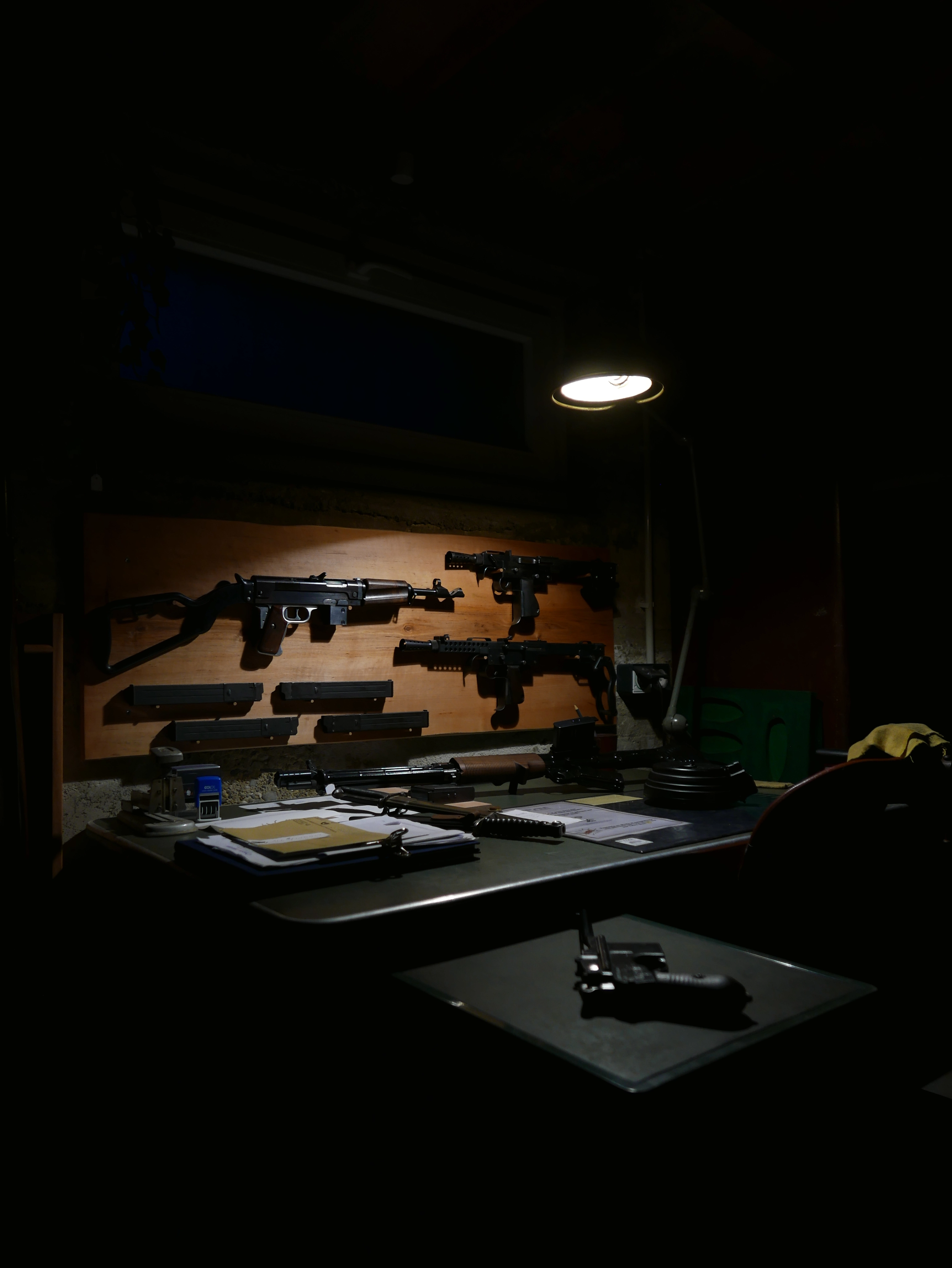 Armes semi-automatiques exposées sur un mur, avec un pistolet et des documents posés sur un établi, sous une lumière tamisée.